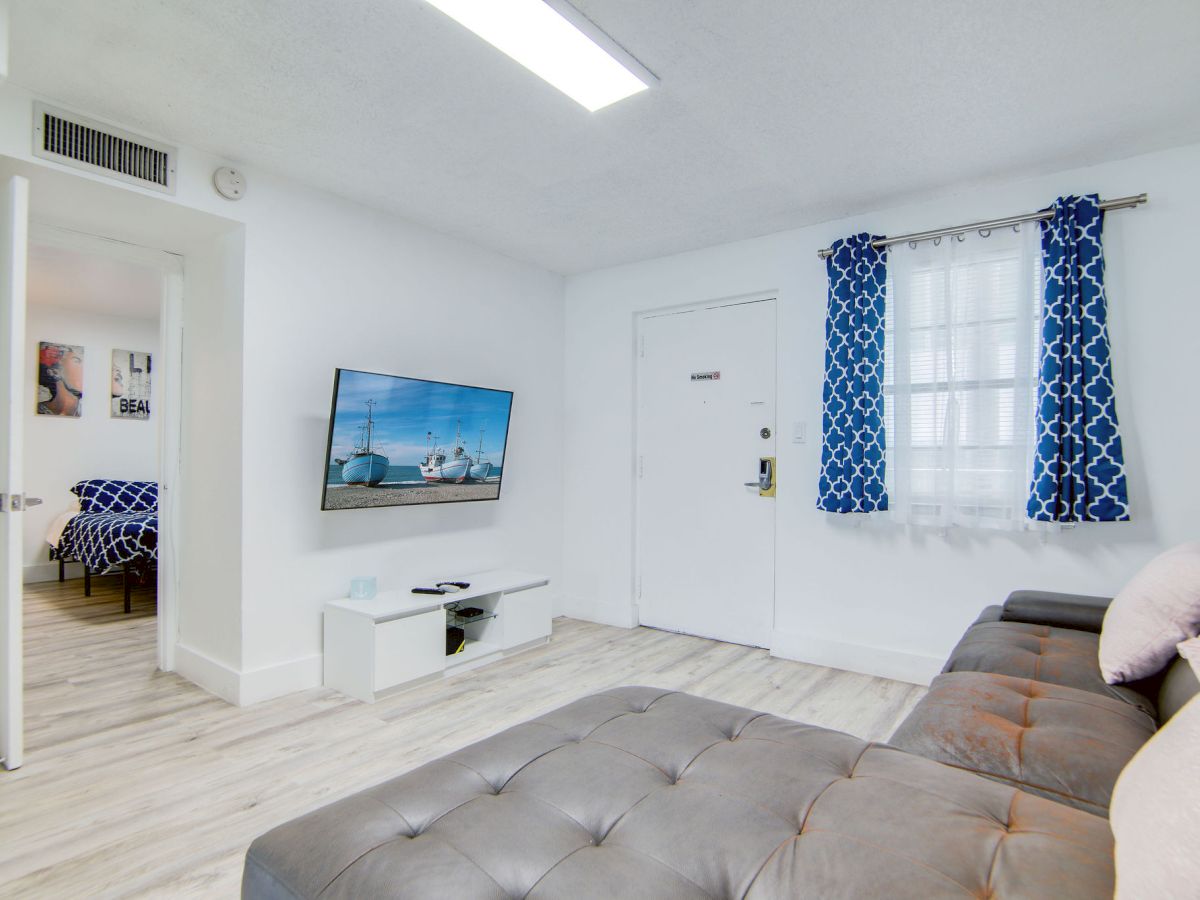 A modern living room with a wall-mounted TV, grey couch, blue patterned curtains, and a view into a bedroom with similar decor.