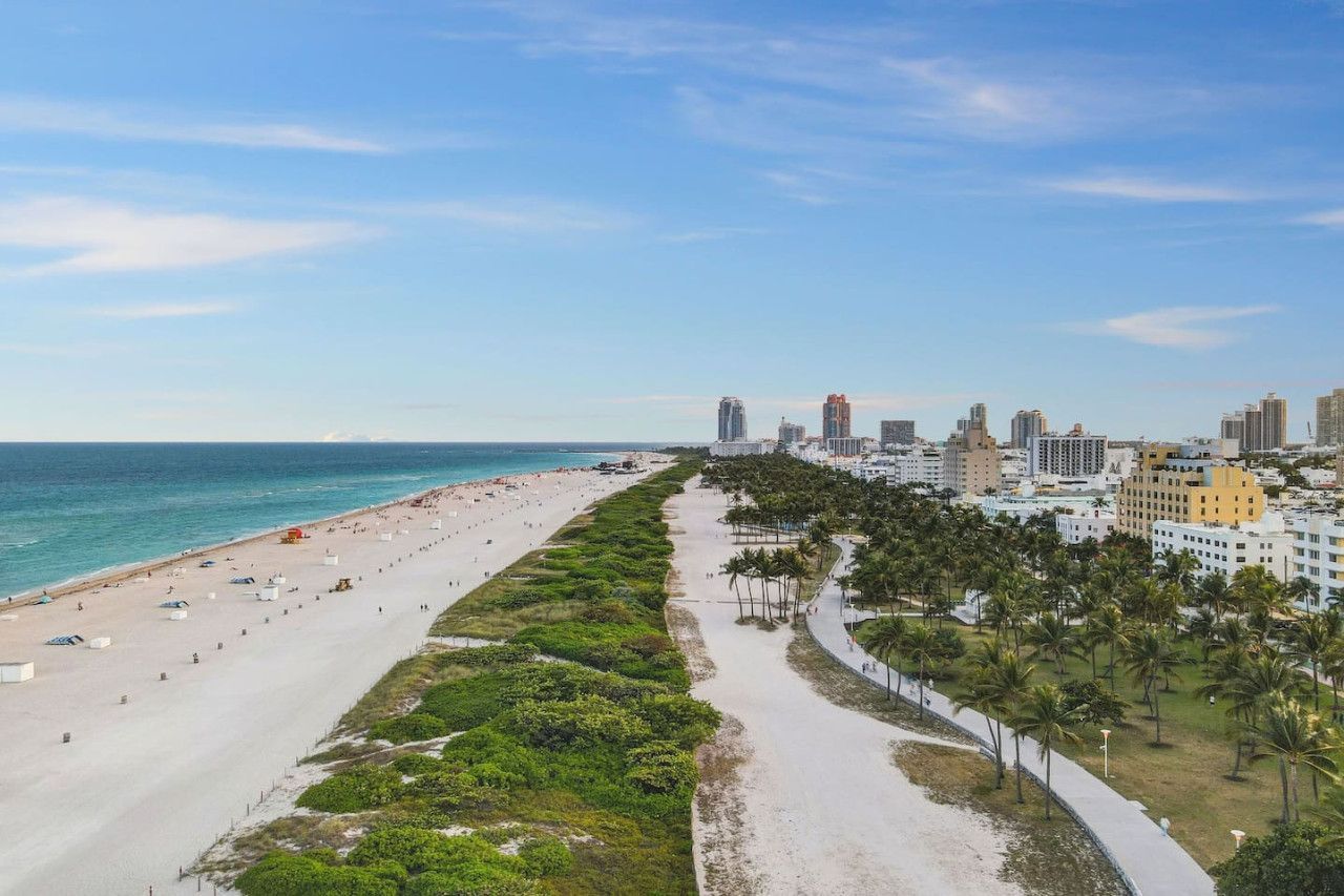 The image shows a coastal city scene with a sandy beach, ocean waves, green areas, a promenade, and buildings in the background.