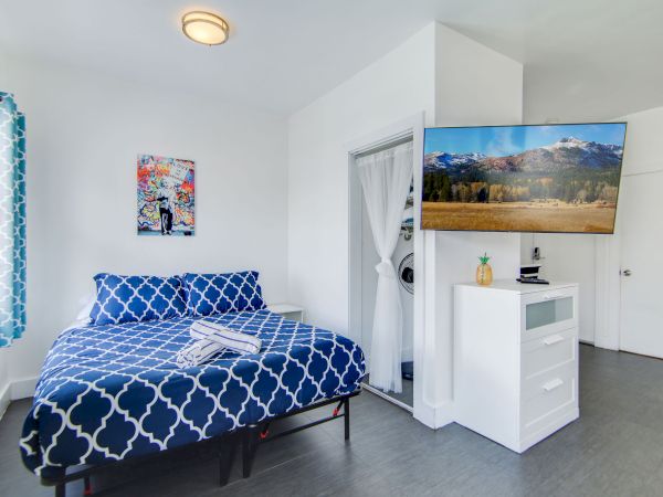 A modern bedroom with a blue patterned bedspread, wall art, a TV on a cabinet, and bright decor in a white room.