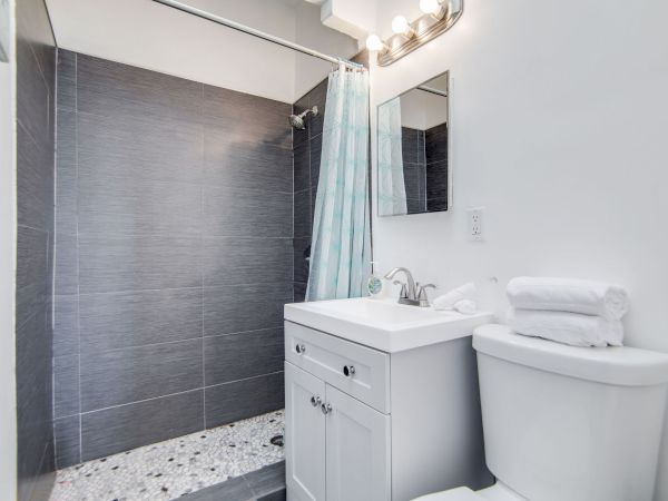 The image shows a small bathroom with a shower, vanity, and toilet. It is white with a blue curtain and neatly folded towels.