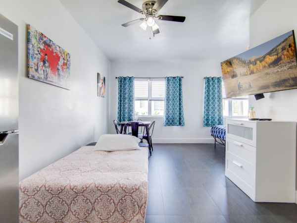 The image shows a bright, modern room with a bed, wall art, blue curtains, a ceiling fan, and a TV mounted above a dresser.
