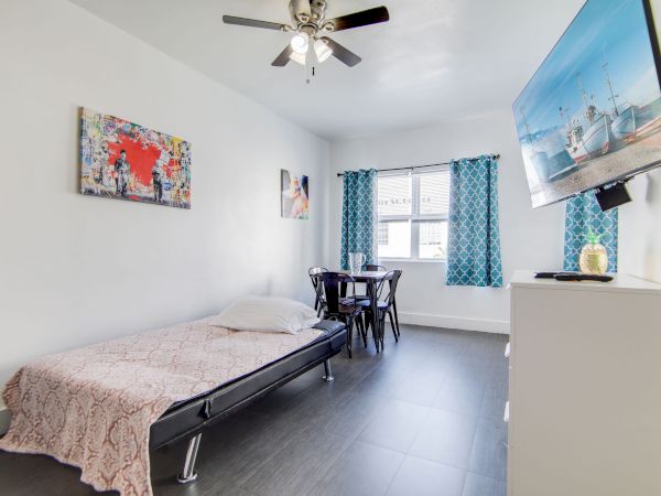 A minimalist room with a twin bed, table, chairs, wall art, fan, TV, and blue curtains in natural light by a window.