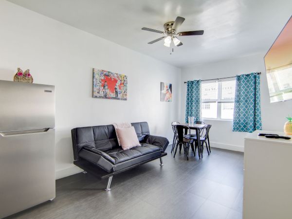 A modern, bright living room with a sofa, dining table, TV, and fridge; decorated with colorful curtains and wall art.