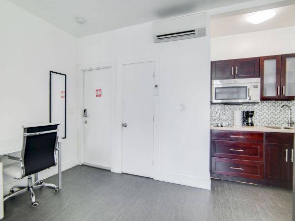 The image shows a small, modern room with a desk and chair, two doors, and a kitchenette with dark cabinets, a microwave, and coffee maker.