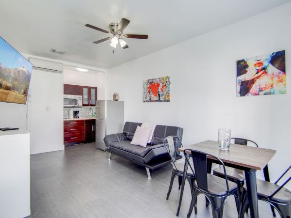 A modern living space with a sofa, dining table, wall art, ceiling fan, and a small kitchen area in the background.