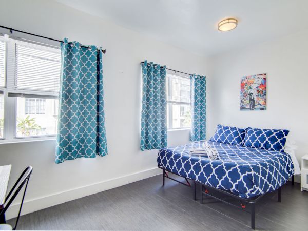 The image shows a bedroom with a bed, blue patterned bedding, matching curtains, a desk, chair, and a piece of artwork on the wall.