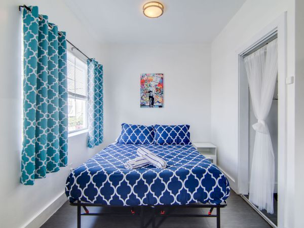 A cozy bedroom with a bed, blue patterned bedding, matching curtains, a small bedside table, artwork on the wall, and natural light.