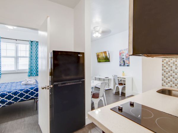 The image shows a small apartment with a kitchenette, adjacent dining area, and a bedroom with a patterned bedspread and blue curtains.