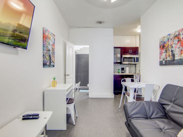A modern, compact living space with a wall-mounted TV, sofa, small dining area, and kitchen, adorned with vibrant artwork on white walls.