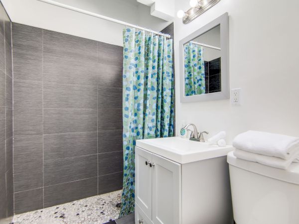 A bathroom with a shower area, blue floral curtain, gray tiles, white vanity, mirror, toilet with towels stacked on top.