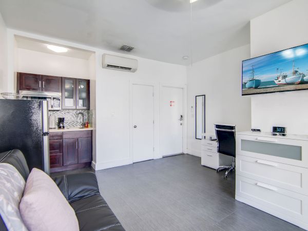 The image shows a small living space with a sofa, a wall-mounted TV, a desk with a chair, and a kitchenette with dark cabinets.