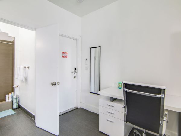 The image shows a minimalist room with a desk, chair, and mirror. A doorway leads to a bathroom with a tub and toiletries visible.