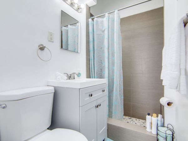 The image shows a small bathroom with a toilet, a vanity with a mirror, and a shower with a blue curtain, neatly organized and bright.