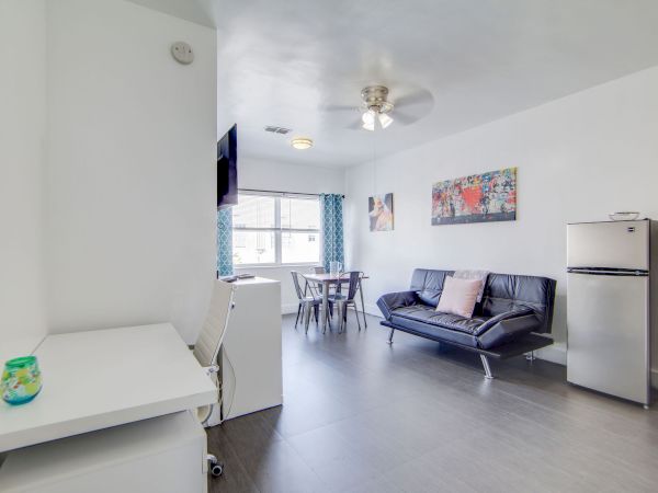 The image shows a modern living space with a sofa, small fridge, table, and chairs. There’s art on the wall and blue curtains by the window.