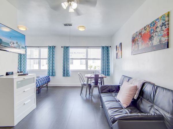 A modern living room with a black sofa, TV, artwork, and a small dining table near three windows with blue curtains ends the sentence.