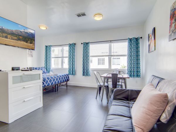 The image shows a bright living area with a TV, bed, table, and chairs, featuring blue curtains and a sofa with pillows for a cozy feel.