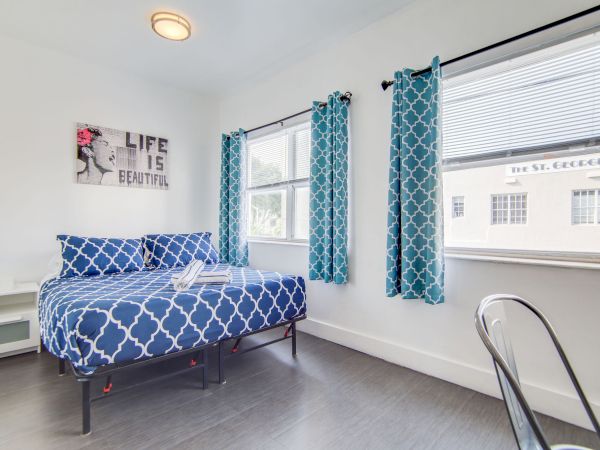 A bright bedroom with a bed dressed in blue patterned bedding, matching curtains, a small desk, and a wall sign saying "Life is Beautiful."