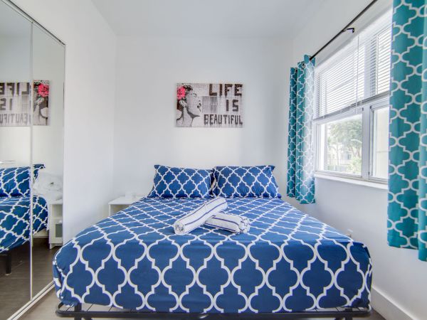 A bedroom with a blue-patterned bedspread, wall art stating "Life is Beautiful," and blue curtains beside a window and mirrored closet doors.
