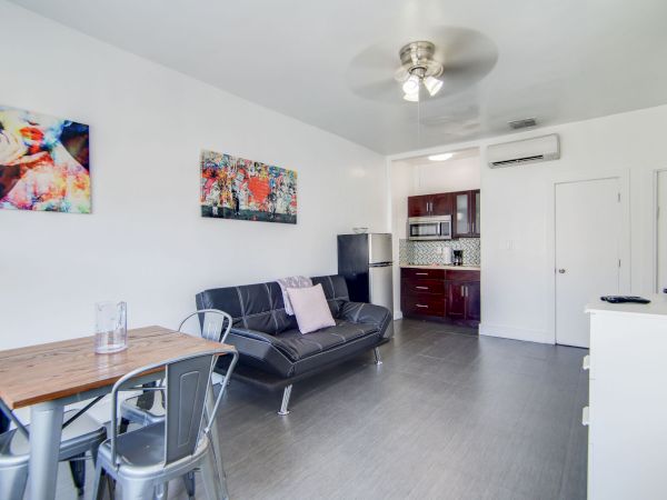 The image shows a modern apartment interior with a small dining table, couch, wall art, kitchenette, and wall-mounted TV.