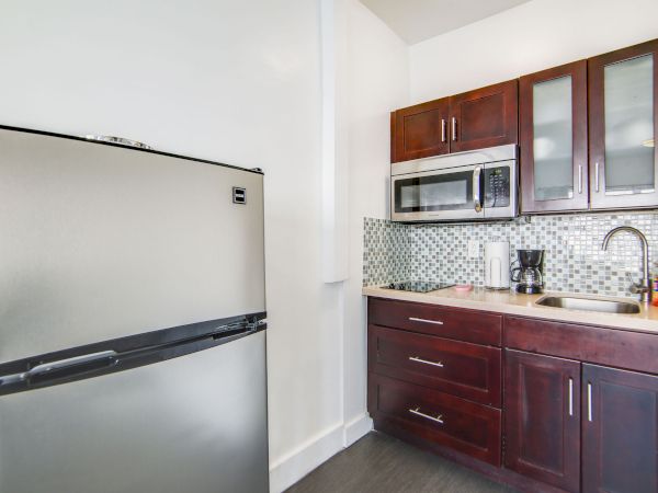 The image shows a compact kitchen with a stainless steel fridge, wooden cabinets, a microwave, a sink, and a tiled backsplash.