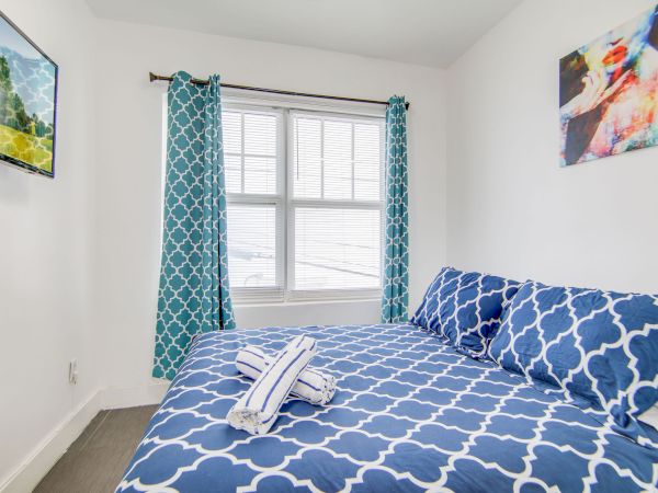 A small bedroom with blue patterned bedding, matching curtains, wall art, a window, and a mounted TV.