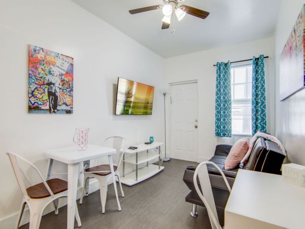 A small living room with a wall-mounted TV, sofa, table, two chairs, vibrant artwork, fan above, and blue patterned curtains at the window.