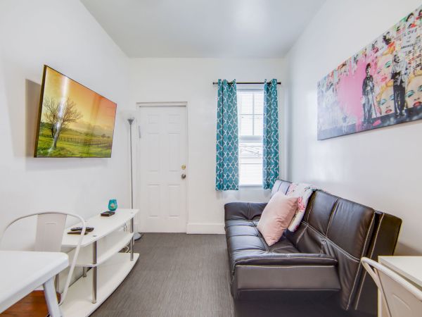 A small living room with a black sofa, wall art, TV, white furniture, and a window with blue curtains next to a white door.