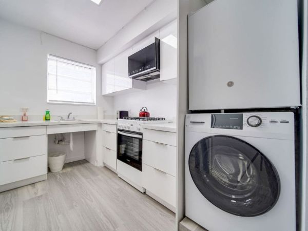 A compact modern kitchen with white cabinets, stove, microwave, and washing machine; bright space with a window and light wood floor.