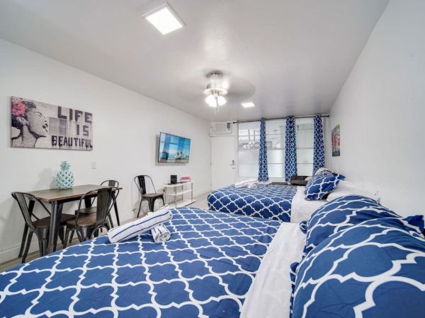 A bedroom with two beds featuring blue patterned bedding, a small dining table, wall art, and a wall-mounted TV, all in a white interior.