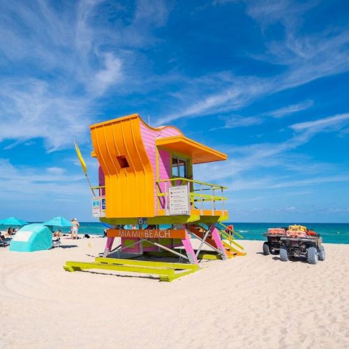 A colorful lifeguard tower stands on a sandy beach with turquoise umbrellas and a clear blue sky in the background.