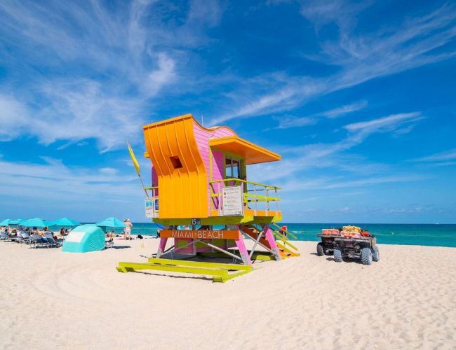 A colorful lifeguard tower stands on a sandy beach with turquoise umbrellas and a clear blue sky in the background.