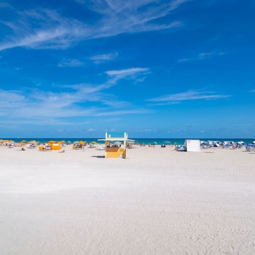 A sunny beach scene with yellow lifeguard stands, beach chairs, and umbrellas under a clear blue sky.