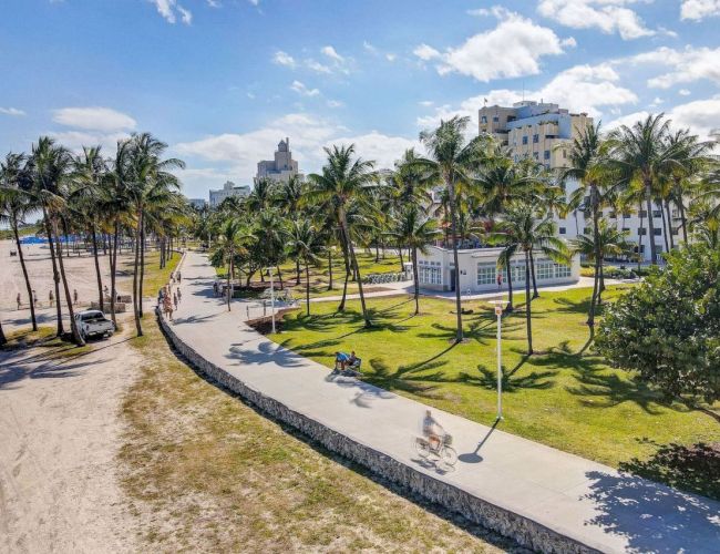 A beachside walkway lined with palm trees and buildings in the background under a blue sky with clouds.