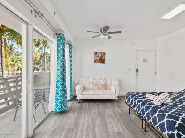 A bright room with a bed, sofa, and balcony overlooking palm trees. There's modern decor, ceiling fan, and blue patterned curtains.