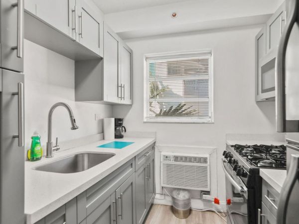 A modern kitchen with gray cabinets, a stainless steel sink, a gas stove, and a window. Cleaning supplies are visible near the sink.