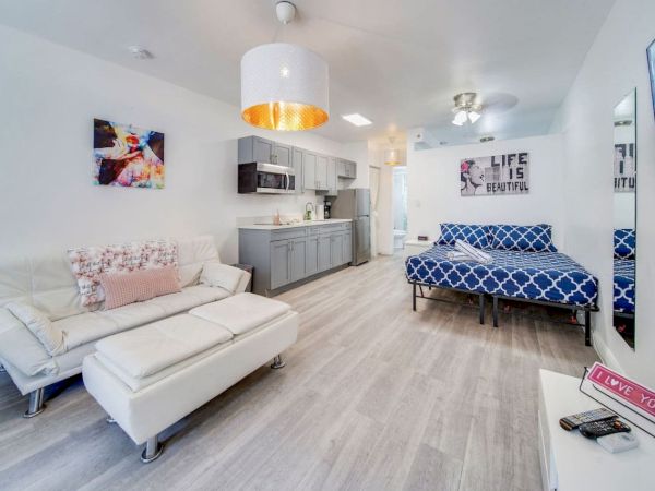 A modern living space with a kitchenette, white sofa, and bed. Decor includes art and a "Life is Beautiful" sign.