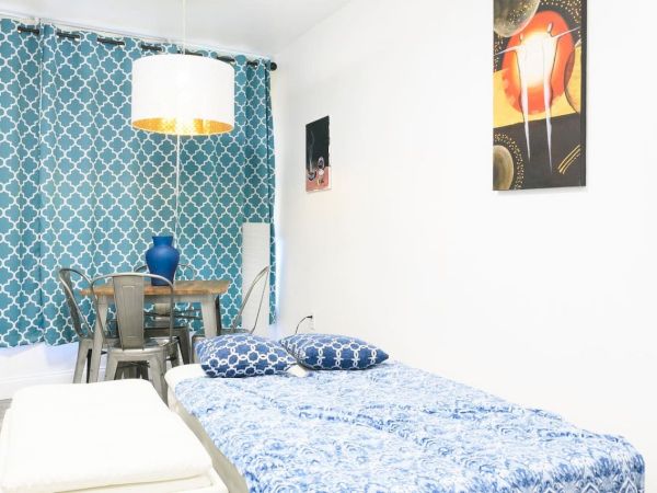 The image shows a room with a bed, a blue patterned curtain, a table with chairs, a blue vase, and wall art, creating a cozy atmosphere.