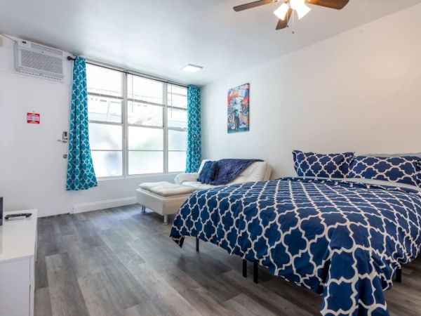 A cozy bedroom with a blue patterned bedspread, large window, white sofa, ceiling fan, and wooden flooring, creating a bright and airy space.