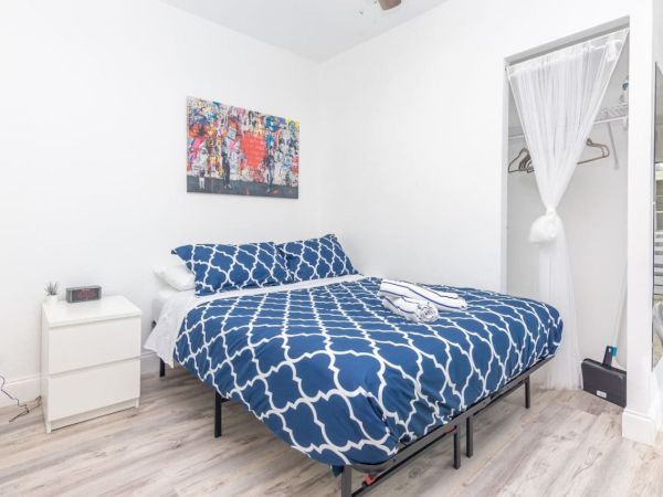 A bedroom with a bed featuring a blue patterned duvet, a small side table, open closet, and colorful wall art, all in a bright space.