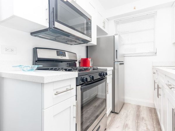 A modern kitchen with white cabinets, stainless steel appliances, a red kettle, and a window with blinds on a light wood floor.