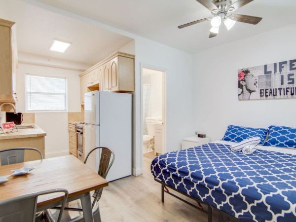 A cozy studio apartment featuring a bed, dining table, compact kitchen, and a bathroom, decorated with a "Life is Beautiful" wall art.