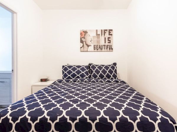 A bedroom features a bed with a geometric-patterned duvet, a bedside table, and wall art saying "Life is Beautiful."