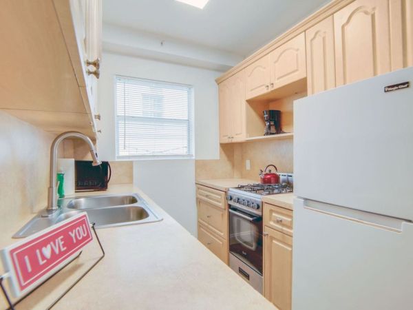 A small kitchen with light wood cabinets, a stove, fridge, and sink. A sign saying "I LOVE YOU" sits on the counter next to the sink.