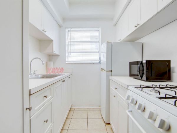 A small kitchen with white cabinets, a stove, fridge, microwave, and a window. Features a pink "XOXO" decoration by the sink.