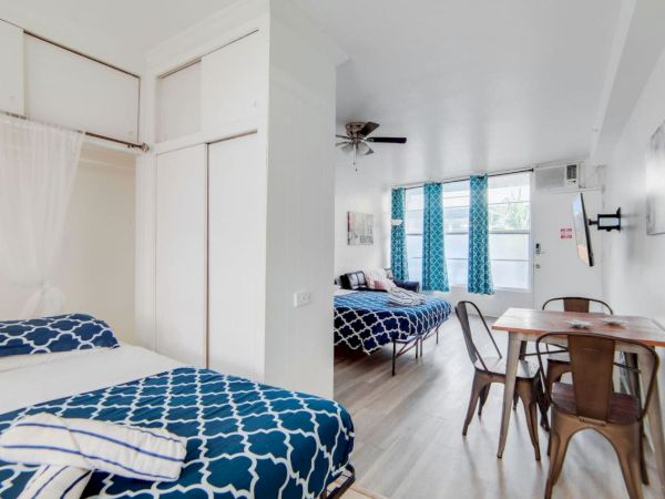 A bright room with two beds, each with blue patterned bedding, a small dining table with chairs, and large windows.