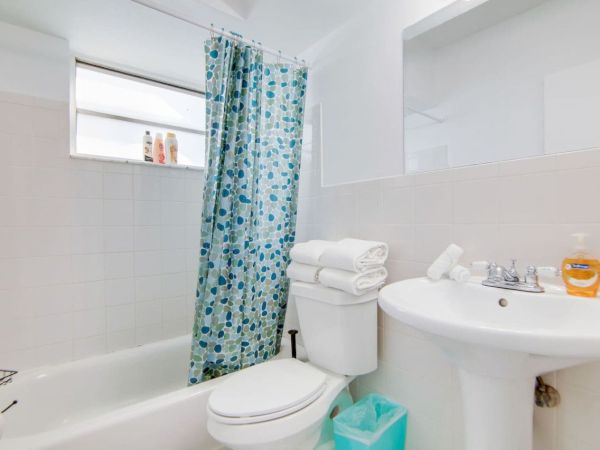 A bathroom with a white tub, blue shower curtain, toilet with towels, pedestal sink with soap, and a small window.