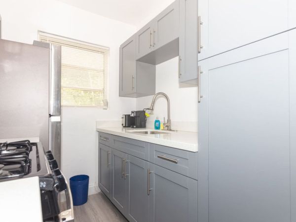 A compact kitchen with light gray cabinets, a stove, a sink, and a small window. A blue trash bin is visible on the floor.