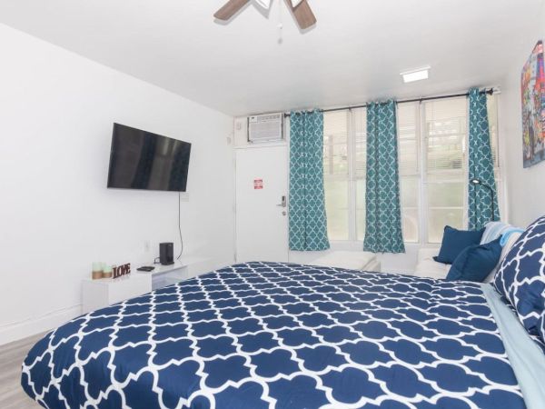 The image shows a bedroom with a blue-patterned bedspread, a wall-mounted TV, curtains, and a door.