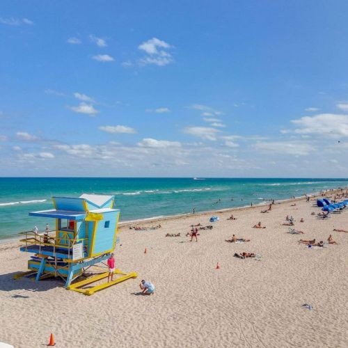 A sunny beach scene with a colorful lifeguard tower, people relaxing on the sand, and a vibrant blue ocean stretching to the horizon.