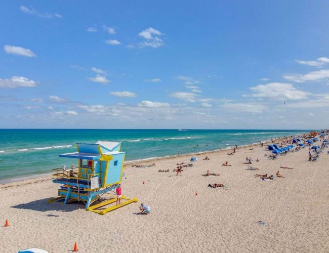 A sunny beach scene with a colorful lifeguard tower, people relaxing on the sand, and a vibrant blue ocean stretching to the horizon.